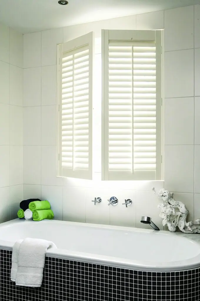 Cửa sổ chớp màu trắng trên bồn tắm có gạch mosaic màu đen tạo điểm nhấn cho không gian. Khăn màu xanh lá cây, xanh nước biển, trắng cuộn tròn đặt trên bồn tắm, tạo cảm giác như một phòng tắm spa.