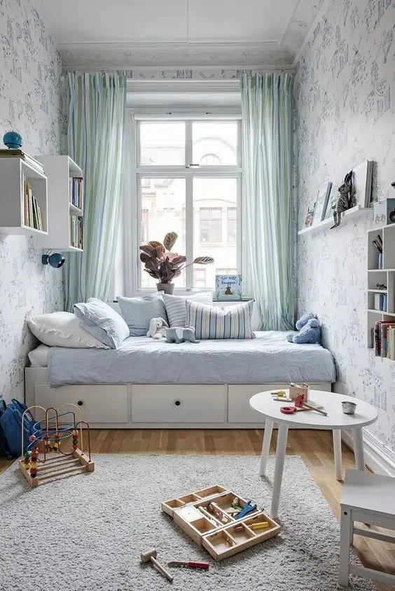Ý tưởng decor phòng ngủ với cách trang trí đơn giản. Thiết kế kê giường gần với cửa sổ, có rèm mỏng để điều chỉnh ánh sáng và đem đến những nét mềm mại hơn cho căn phòng. Kệ gỗ treo, hộc tủ treo tường kết hợp với giường thông minh chứa ngăn kéo đựng quần áo phía dưới sẽ giúp giải phóng không gian nhỏ của căn phòng.